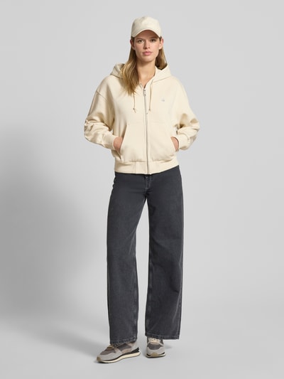 Kobiece modelka nosi bluzę rozpinaną z kapturem i kieszenią kangurka od Carhartt Work In Progress, bawełna, krój relaxed fit, zamek błyskawiczny, wyhaftowane logo, widok całej sylwetki z przodu.