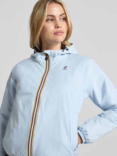 Modelka nosi jasnoniebieską Regular Fit Jacke mit Logo-Print Modell 'CLAUDETTE' od K-WAY; elastyczne mankiety, kaptur ze ściągaczem, szerokość w rozmiarze S: 51 cm, długość: 59 cm, szerokość ramion: 39 cm, długość rękawa: 63 cm, zdjęcie z przodu, zbliżenie na górną część ciała.