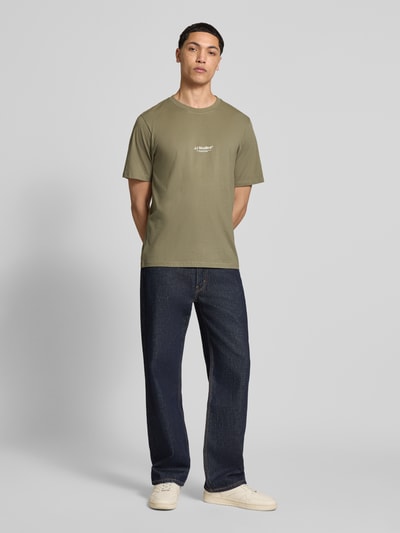 Męski model nosi T-Shirt mit Logo und Rundhalsausschnitt von Jack & Jones z bawełny ekologicznej, krój regular fit, logo, długość tyłu 68 cm, kolor khaki, gładka faktura, całopostaciowy widok z przodu.
