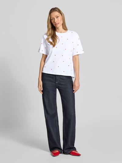 Frauenmodel trägt Boxy Fit T-Shirt aus reiner Baumwolle von Tom Tailor Denim, weiß mit roten Herzstickereien, unifarben, Rundhals, kurzarm, dunkle Jeans, rote Schuhe, ganzkörperansicht von vorne.
