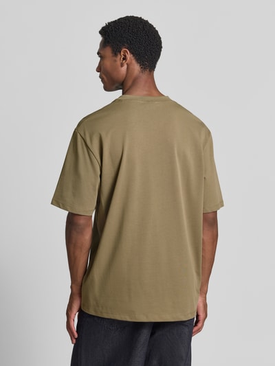 Męski model nosi Oversized T-Shirt aus reiner Baumwolle von Lindbergh, kolor oliwkowy, miękka faktura, długość tyłu w rozmiarze M: 70 cm, krój oversized, czysta bawełna, lekko opadające ramiona, półrękawkowy design, Ganzkörperansicht Rückansicht.