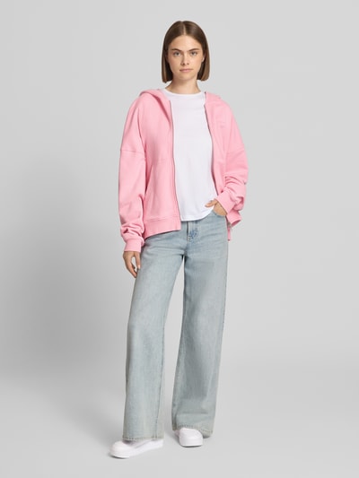 Kobiece model prezentuje Oversized Sweatjacke mit Zweiwege-Reißverschluss Modell 'Flower' von OH APRIL z bawełny, krój oversized, bluza z kapturem, kieszeń kangurka, szerokie rękawy, jasnoróżowy odcień, gładka tekstura, widok całej sylwetki od przodu.