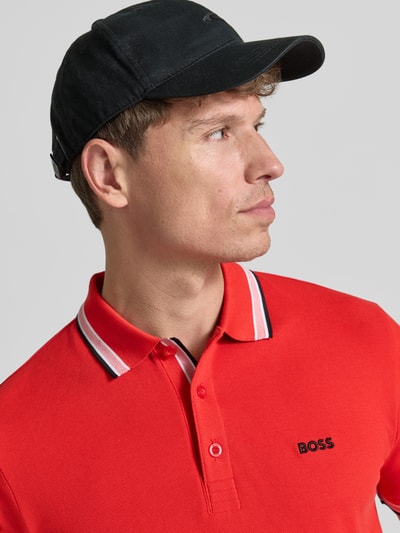 Mężczyzna nosi koszulkę polo Regular Fit Poloshirt aus reiner Baumwolle model 'PADDY' od BOSS Green w kolorze czerwonym, teksturowana bawełna, detaliczne kontrastowe wykończenia przy kołnierzyku, zbliżenie na górną część ciała