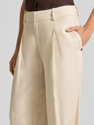 Kobieta nosi Wide Leg Pants aus Viskose-Mix mit Biesen Modell 'MIssa Coleen' od MOS MOSH w kolorze jasnobeżowym, tekstura lniana, loose fit, kieszenie z guzikiem, kontekst: damskie spodnie wide leg, mieszanka wiskozy z domieszką lnu, szerokość w rozmiarze 36: 39 cm, długość wewnętrzna nogawki: 106 cm, zapięcie na haczyk i zamek, biesy, zbliżenie na górną część przodu.