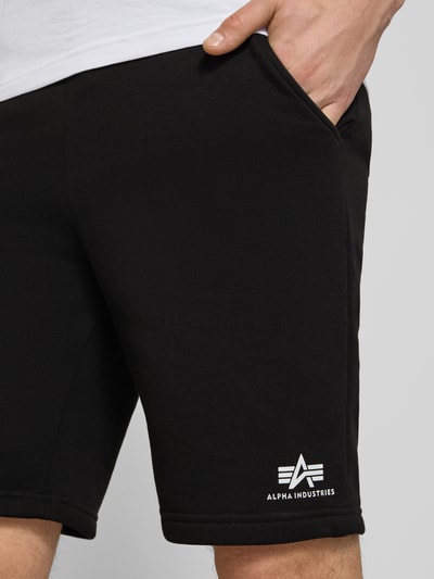 Männermodel trägt Shorts mit Eingrifftaschen und Logo-Print von Alpha Industries in Schwarz aus Baumwoll-Mix, Nahaufnahme Oberschenkel, Relaxed-Fit, elastischer Bund mit Kordelzug, seitliche Eingrifftaschen, Logo-Print, Gesäßtasche, Bundweite bei Größe M 39 cm, Innenbeinlänge 24 cm.