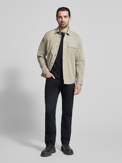Mężczyzna nosi Regular Fit Hemdjacke mit Brusttaschen od camel active, mieszanka bawełny z dodatkiem streczu, kołnierzyk typu kent, listwa guzikowa, kieszenie na piersi, długość tyłu w rozmiarze M 74 cm, szerokość ramion w rozmiarze M 47 cm, szerokość klatki piersiowej w rozmiarze M 58 cm, długość rękawów w rozmiarze M 66 cm, jasny beżowy kolor, kontrast z czarnymi spodniami, pełen widok sylwetki z przodu.