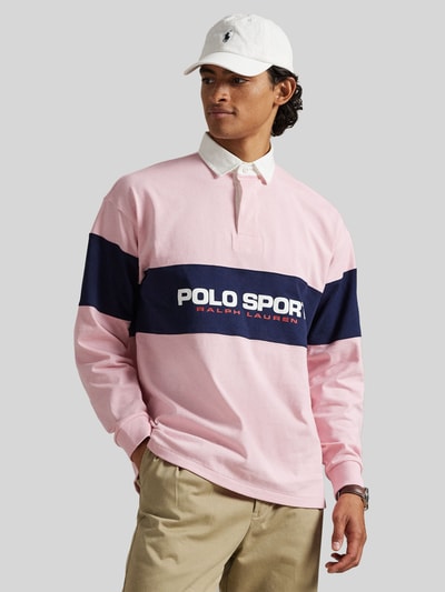 Männermodel trägt Regular Fit Poloshirt aus reiner Baumwolle von Polo Ralph Lauren in Rosa mit navyfarbenem Streifen und Label-Schriftzug, Ganzkörperansicht von vorne.