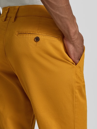 Męski model nosi Hose mit Gesäßtaschen od Christian Berg Men, bawełna-elastan, krój slim fit, jasnopomarańczowy kolor, kieszenie z tyłu, szerokość w rozmiarze 50: 45 cm, długość nogawki od wewnątrz: 80 cm, zbliżenie na biodra i kieszeń.