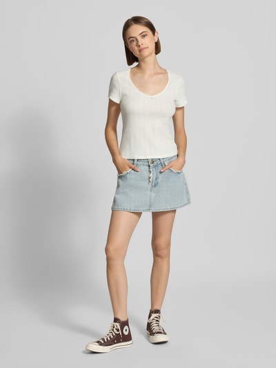 Modelka kobieca nosi biały damski t-shirt z krótkim rękawem model 'SWEETIE' od Levi's®, jasna dżinsowa spódnica, brązowe trampki, jasnoszare tło, damski t-shirt od Levi's®, pełny widok z przodu.