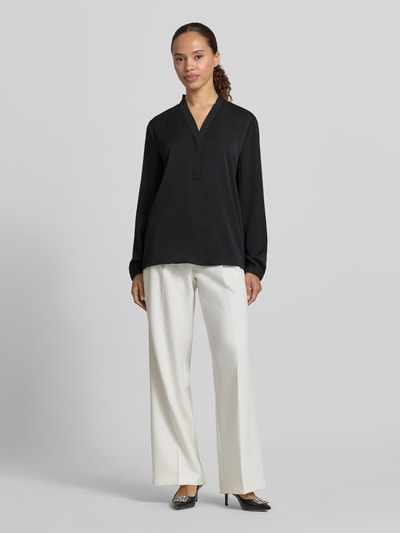 Frauenmodel trägt Bluse mit V-Ausschnitt und Label-Detail von Marc Cain, Loose-Fit, schwarz, elastische Ärmelabschlüsse, Ganzkörperansicht von vorne. Rückenlänge bei Größe 36: 46 cm, Ärmellänge bei Größe 36: 60 cm.