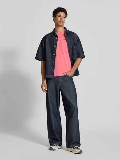 Männermodel trägt Regular Fit T-Shirt mit Logo-Stitching von Superdry in Pink, kombiniert mit weiter Jeans und Sneakers, Ganzkörperansicht von vorne.