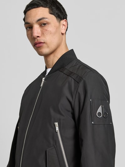 Männermodel trägt Bomberjacke mit Zweiwege-Reißverschluss von Moose Knuckles in Schwarz mit Stehkragen, Ärmeltaschen, Nahaufnahme Oberkörper. Rückenlänge 67 cm, Schulterbreite 46 cm, Ärmellänge 68 cm, lockere Passform, elastische Abschlüsse.