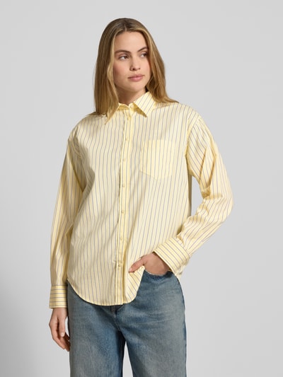 Frauenmodel trägt Relaxed Fit Bluse mit Brusttasche von Gant aus reiner Baumwolle, gestreiftes Gelb mit blauen Linien, Ganzkörperansicht von vorne.