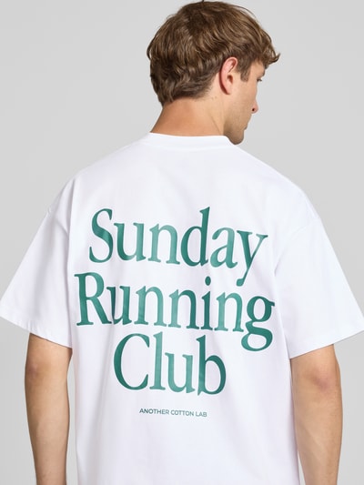 Męski model nosi T-Shirt mit Print und Rundhalsausschnitt od ANOTHER COTTON LAB, biała bawełna, krój relaxed fit, nadruk, długość tyłu 69 cm w rozmiarze M, zdjęcie całej sylwetki z tyłu.