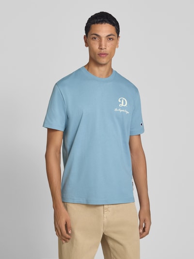 Männermodel trägt Relaxed Fit T-Shirt aus reiner Baumwolle von Champion in Hellblau mit Statement-Print, Ganzkörperansicht von vorne. Herren T-Shirt, reine Baumwolle, Relaxed Fit, Label-Stitching, Rückenlänge bei Größe M 70 cm.