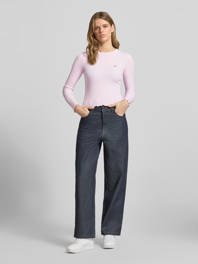 Modelka nosi Slim Fit Langarmshirt aus Baumwoll-Mix von Tommy Jeans w paski, detal z logo, okrągły dekolt, drobne prążkowanie, mieszanka bawełny, długi rękaw, zakończenia z falbanką, widok całej sylwetki z przodu.
