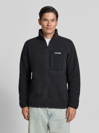 Männermodel trägt Fleecejacke mit Stehkragen Modell 'Jasper' von Columbia in Schwarz mit Reißverschlusstaschen, Ganzkörperansicht von vorne. Rückenlänge, Ärmellänge und Schulterbreite in Größe M entsprechen dem Kontext.