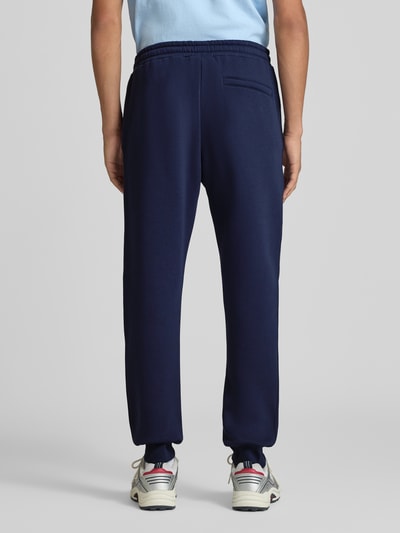 Męski model nosi Slim Fit Sweatpants aus Baumwoll-Mix von REVIEW w kolorze granatowym, wpuszczane kieszenie i kieszeń z tyłu, wyhaftowane logo, krój slim fit, elastyczny pas z tunelem, mieszanka bawełny, długość wewnętrzna nogawki w rozmiarze M: 79 cm, widok całej sylwetki z tyłu.