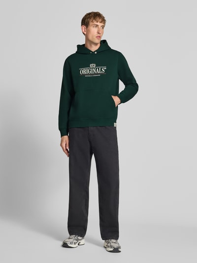 Männermodel trägt Hoodie mit Label-Print Modell 'FREDERIKSBERG' von JACK & JONES, dunkelgrün, lockerer Schnitt, Ganzkörperansicht von vorne. Baumwoll-Mix, Kapuze, Kängurutasche, gerippte Abschlüsse, Rückenlänge M 65 cm, Ärmellänge M 67 cm.