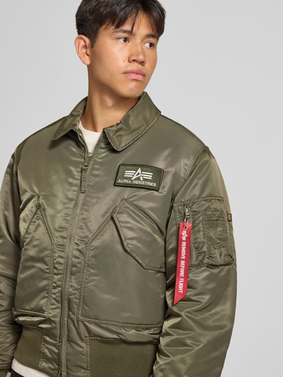 Männermodel trägt Bomberjacke mit seitlichen Pattentaschen und Label-Patch von Alpha Industries in Olivgrün, Nahaufnahme Oberkörper.