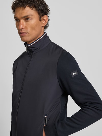 Männermodel trägt Sweatjacke mit Stehkragen von bugatti aus dunkelblauem Baumwoll-Mix, Label-Detail, Reißverschlusstaschen, Nahaufnahme Oberkörper.