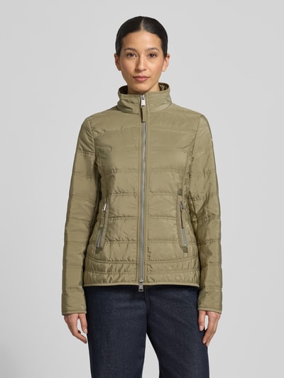 Kobiece model prezentuje kurtka pikowana z stójką i kieszeniami zapinanymi na zamek błyskawiczny 'Steppjacke mit Stehkragen und Reißverschlusstaschen' od Fuchs Schmitt, taliowany krój, detal z logo, zamek błyskawiczny dwustronny, zielony kolor, długość tyłu w rozmiarze 36: 65 cm, szerokość ramion w rozmiarze 36: 45 cm, szerokość klatki piersiowej w rozmiarze 36: 42 cm, długość rękawów w rozmiarze 36: 65 cm, widok całej sylwetki z przodu.