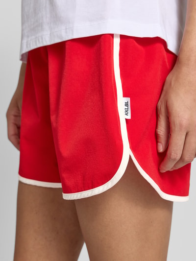 Frauenmodel trägt Track Shorts mit elastischem Bund und Label-Detail von Karo Kauer in Rot mit weißem Saum, Material-Mix mit Stretch-Anteil, Loose-Fit, Nahaufnahme Oberschenkel.