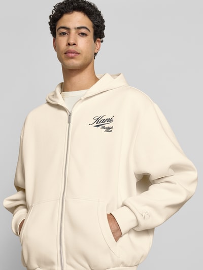 Männermodel trägt Oversized Sweatjacke mit Logo-Stitching von KARL KANI in Creme, Kängurutasche, Baumwoll-Mix, Nahaufnahme Oberkörper.