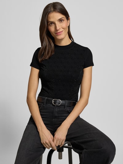Kobiece model prezentuje Slim Fit T-Shirt mit Allover-Strasssteinbesatz od Guess, czarny, bawełna i elastan, stójka, detal z logo, długość tyłu 51 cm, obszycie z kamieni stras na całej powierzchni, dopasowany krój, zbliżenie na górną część ciała.