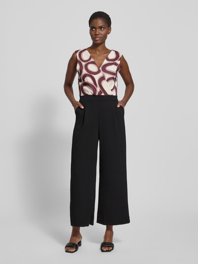Frauenmodel trägt Jumpsuit mit Eingrifftaschen Modell 'Bi44lina' von ZABAIONE aus Material-Mix mit Stretch-Anteil, weites Bein, V-Ausschnitt, Ganzkörperansicht von vorne. Rückenlänge bei Größe S 40 cm, Brustweite 35 cm, Gesamtlänge 140 cm, Innenbeinlänge 67 cm. Geometrisches Muster in Creme und Bordeaux am Oberteil, Hose in Schwarz, glatte Textur.