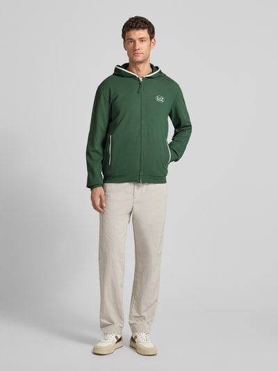 Mężczyzna-model nosi zieloną Sweatjacke mit Logo und Zweiwege-Reißverschluss od EA7 Emporio Armani z kapturem i kieszeniami, widok całej sylwetki z przodu.