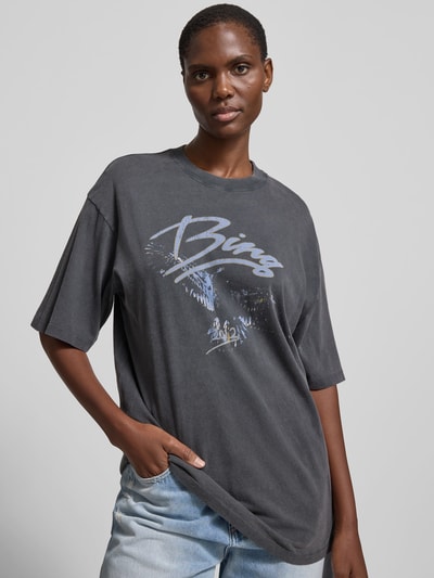 Frauenmodel trägt Oversized T-Shirt mit Logo-Print von Anine Bing aus Reiner Baumwolle, Oversized-Fit, Rundhalsausschnitt, Halbarm, dunkle Farbe, dezentes Logo-Print-Design, Rückenlänge bei Größe S 70 cm, Ganzkörperansicht von vorne.