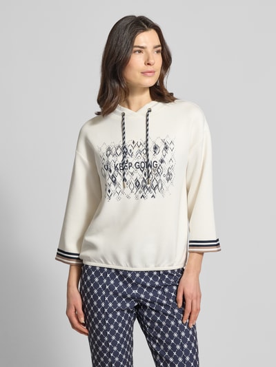 Modelka nosi bluzę 'Sweatshirt aus Viskose-Mix mit 3/4-Arm' od Betty Barclay, mieszanka wiskozy z dodatkiem streczu, krój relaxed fit, nadruk z napisem, kaptur z tunelem, rękaw o dł. 3/4, jasne kolory, proste kształty, wyraźne tekstury. Całkowity widok z przodu.