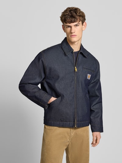 Männermodel trägt Jeansjacke mit Brusttasche und Label-Detail von Carhartt Work In Progress, dunkles Denim, Reißverschluss, Regular Fit, Ganzkörperansicht von vorne