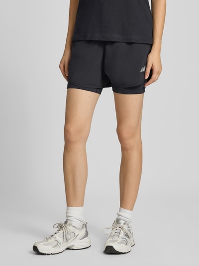 Frauenmodel trägt schwarze Shorts im 2-in-1-Look mit Logo von New Balance, elastischer Bund, Regular Fit, Nahaufnahme Unterkörper.