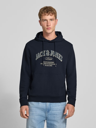 Männermodel trägt dunkelblauen Regular Fit Hoodie aus Baumwoll-Mix Modell 'Denver' von JACK & JONES mit Logo, Känguru-Tasche, Ganzkörperansicht von vorne.