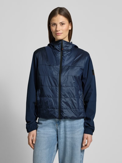 Frauenmodel trägt dunkelblaue Jacke mit Zweiwege-Reißverschluss Modell 'MARTA' von FIRE + ICE, leichte Struktur, Ganzkörperansicht von vorne. Damenjacke Regular Fit, Kapuze mit Tunnelzug, Rückenlänge 57 cm bei Größe 36, dezente Steppnähte, glatte Oberfläche.