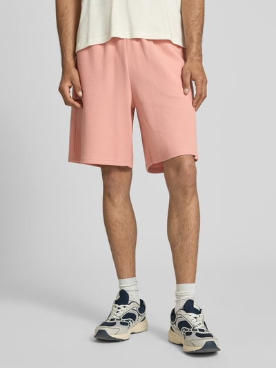 Männermodel trägt Regular Fit Shorts mit Strukturmuster von KARL KANI aus Baumwoll-Mix in Rosa mit Logo-Stitching, elastischer Bund, Ganzkörperansicht von vorne