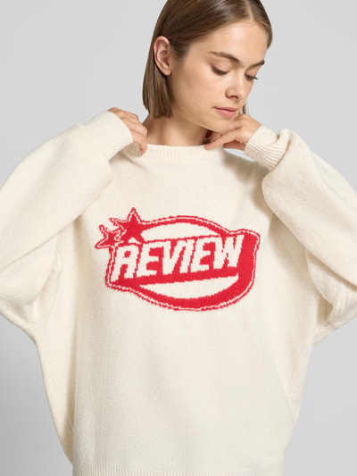 Frauenmodel trägt Strickpullover mit Label-Print von REVIEW in Weiß mit rotem Print, lockerer Schnitt, Rundhalsausschnitt, Nahaufnahme Oberkörper.