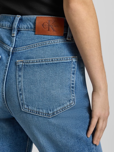 Männermodel trägt Straight Fit Jeans mit Logo-Patch Modell 'Mom' von Calvin Klein Jeans, 5-kieszeniowy design, koniakowa naszywka z logo, niebieski denim, zapięcie na guzik i zamek, szerokość w rozmiarze 27/32: 36 cm, długość wewnętrzna nogawki: 75 cm, detailaufnahme.