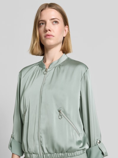 Kobiece modelka nosi bluzon z satyny o kroju regular fit, stójka, zamek błyskawiczny, kieszenie zapinane na zamek błyskawiczny, regulowana długość rękawów, Blouson mit regulierbarer Ärmellänge und Reißverschlusstaschen od Betty Barclay, długość tyłu w rozmiarze 36: 53 cm, szerokość ramion: 38 cm, szerokość w rozmiarze 36: 50 cm, długość rękawów w rozmiarze 36: 57 cm, jasny odcień zieleni, gładka błyszcząca struktura, widok na górną część ciała z przodu.