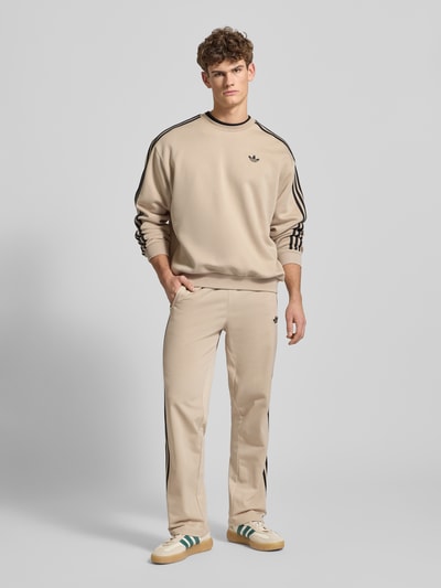 Männermodel trägt beige Heren Trainingshose met logoprint, ritszakken, elastische band, model 'FIREBIRD' van adidas Originals, regular fit, ganzkörperansicht von vorne.