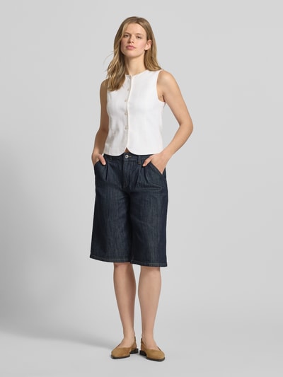 Kobieta modelka nosi Long Shorts z wpuszczanymi kieszeniami od Gina Tricot, mieszanka bawełny z dodatkiem streczu, krój loose fit, zapięcie na guzik i zamek błyskawiczny, wpuszczane kieszenie boczne i kieszenie z tyłu, szerokość w rozmiarze 36: 42 cm, długość wewnętrzna nogawki w rozmiarze 36: 31 cm, ciemny denim, gładka tekstura, prosty fason, jasna bluzka, całkowity widok z przodu.