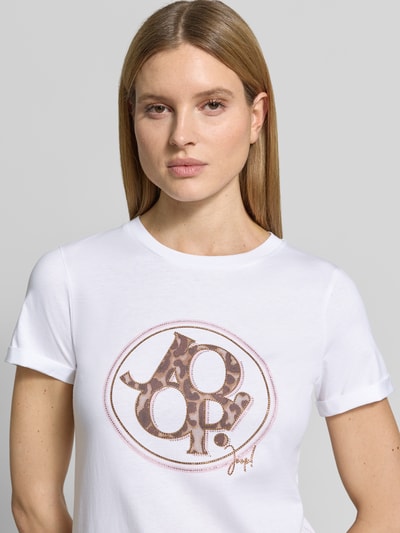Kobieta nosi T-shirt z obszyciem z kamieni stras, logo oraz detal z logo, damski t-shirt z bawełny od Joop!, prosty krój, okrągły dekolt, długość tyłu w rozmiarze 36: 59 cm, zbliżenie na górną część ciała