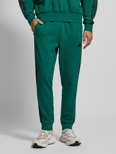 Männermodel trägt Sweatpants mit elastischem Bund von adidas Sportswear im Baumwoll-Mix mit Viskose-Anteil, Lockerer Schnitt, Tunnelzug, Label-Stitching, Farbe grün mit schwarzen Details, mittlere Struktur, Ganzkörperansicht von vorne.