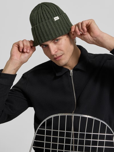 Männermodel trägt Beanie aus Woll-Mix von Forét in Dunkelgrün, Ripp-Optik, breiter Umschlag, Label-Patch, Logo-Schriftzug, Nahaufnahme Oberkörper