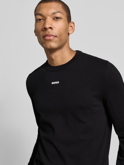 Mężczyznamodel ma na sobie Regular Fit Strickpullover aus reine Baumwolle Modell 'SAN CASSIUS-C2' od HUGO, krój regular fit, kolor czarny, crew neck, długi rękaw, prążkowane wykończenia, dzianina, nadruk logo, unikatowa tekstura bawełny, materiał naturalny. Ujęcie: z przodu, zbliżenie na górną część ciała.