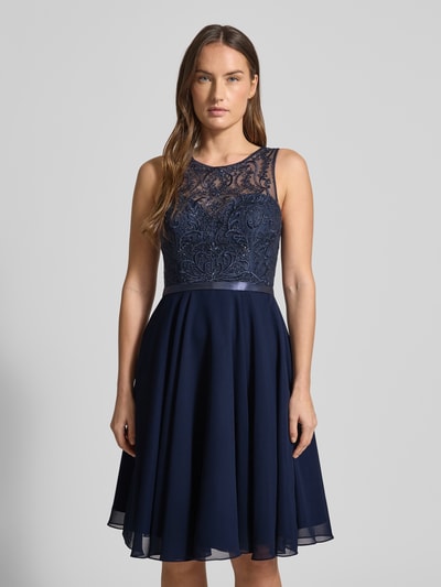 Kobiece model nosi Sukienka koktajlowa z obszyciem z kamieni stras od LUXUAR FASHION, materiał wytrzymały, taliowany krój, długość do kolan, jednokolorowy, zamek błyskawiczny, okrągły dekolt, półprzezroczysty materiał, długość tyłu 96 cm, szerokość klatki piersiowej 41 cm, widok całej sylwetki z przodu.