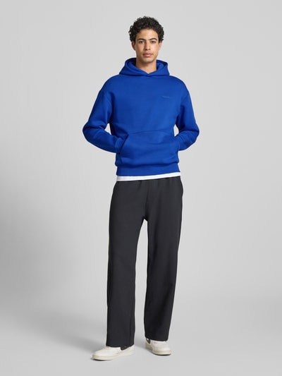 Männermodel trägt Hoodie mit Känguru-Tasche von REVIEW in Blau, Baumwollmischung, lockere Passform, Kapuze, Label-Detail, elastische Abschlüsse, Ganzkörperansicht von vorne.