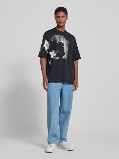 Männermodel trägt Comfort Fit T-Shirt mit Motiv-Print von Armani Exchange, Allover-Look in Schwarz mit Rundhalsausschnitt, Herren T-Shirt, Rückenlänge 76 cm bei Größe M, Ganzkörperansicht von vorne.
